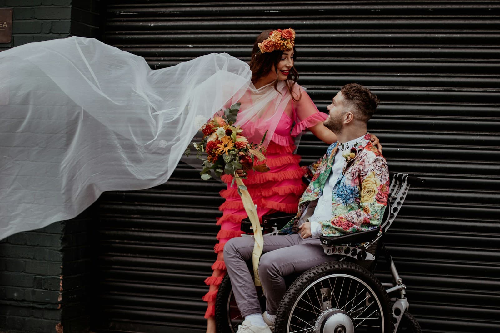 Een stel voor een donker rolluik: zij in een felroze jurk met een wapperende sluier, hij in een rolstoel in een jasje met bloemen; ze lachen naar elkaar